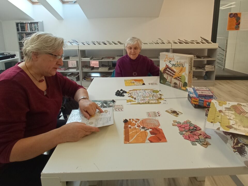 Seniorka czyta instrukcję do gry "Zamek Smoków", druga seniorka słucha