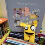Żółty stworek z filcu i w tle książka - Kto mieszka za meblami