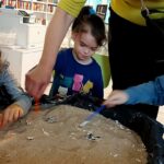 Dzieci wygrzebują w piasku kości dinozaurów z patyków