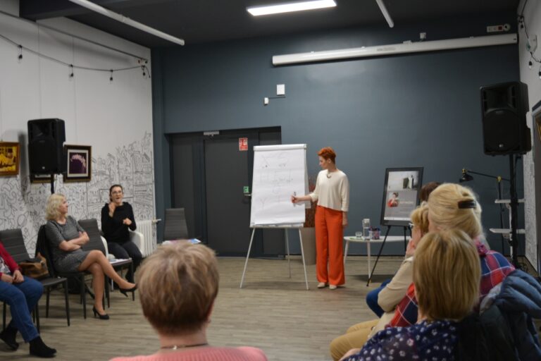 Aleksandra Frątczak stoi koło tablicy i tłumaczy, goście słuchają