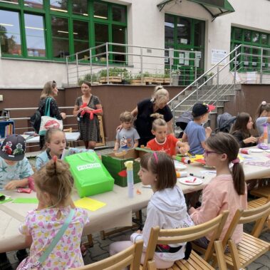 Na zewnątrz biblioteki, przy stole dzieci wykonują prace plastyczne.