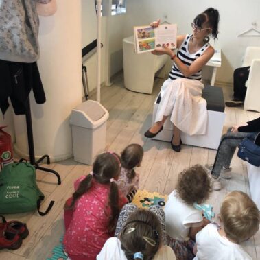 Dzieci siedzą na piankowym dywanie w głębi bibliotekarka głośno czyta i pokazuje ilustracje