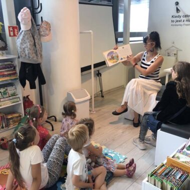 Dzieci siedzą na piankowym dywanie w głębi bibliotekarka głośno czyta i pokazuje ilustracje