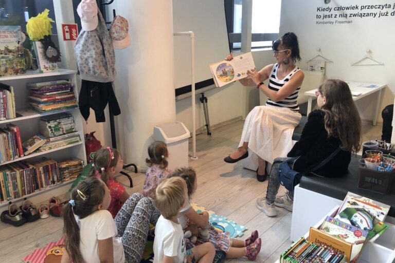 Dzieci siedzą na piankowym dywanie w głębi bibliotekarka głośno czyta i pokazuje ilustracje