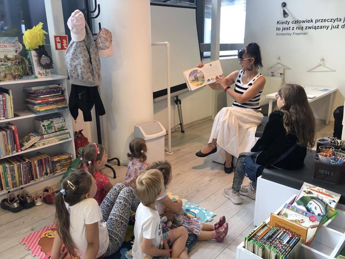 Dzieci siedzą na piankowym dywanie w głębi bibliotekarka głośno czyta i pokazuje ilustracje