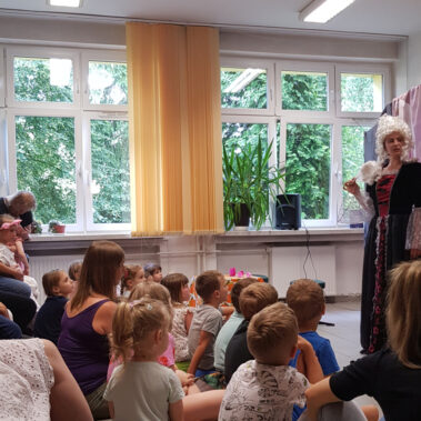 Dzieci siedzą na podłodze i słuchają przestawienia, a na scenie grają dwie aktorki