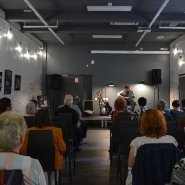 Widok na salę z sceną, na której siedzi Zygmunt i gra na gitarze