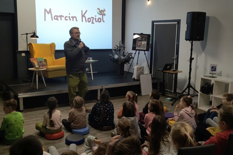 Marcin Kozioł przemawia, dzieci siedzą na pufkach i krzesłach
