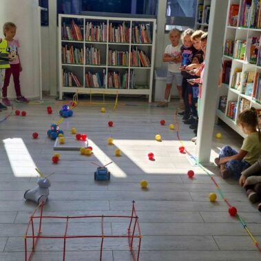 Dwie drużyny rywalizują sterując robotami. Po podłodze roboty poruszają pchając kulki.