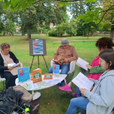 Przy stoliku siedzą czytelnicy w towarzystwie bibliotekarki i czytają na głos książki