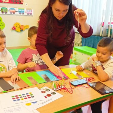 Bibliotekarka pokazuje jak grać na Makey Makey z użyciem kartek z naklejonymi foliami, chłopcy oglądają