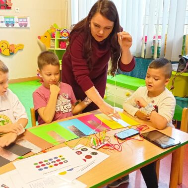 Bibliotekarka pokazuje jak grać na Makey Makey z użyciem kartek z naklejonymi foliami, chłopcy oglądają