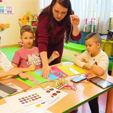 Bibliotekarka pokazuje jak grać na Makey Makey z użyciem kartek z naklejonymi foliami, chłopcy oglądają i zaczynają grać