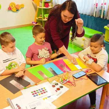 Bibliotekarka pokazuje jak grać na Makey Makey z użyciem kartek z naklejonymi foliami, chłopcy oglądają