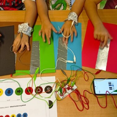 Ręce grają dotykając folie. Obok podłączony jest moduł Makey Makey do folii i podłączony do smartfona