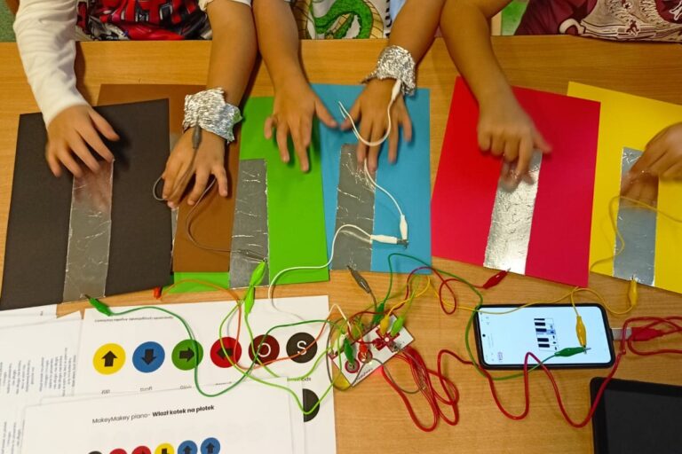 Ręce grają dotykając folie. Obok podłączony jest moduł Makey Makey do folii i podłączony do smartfona