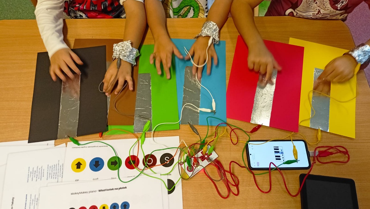 Ręce grają dotykając folie. Obok podłączony jest moduł Makey Makey do folii i podłączony do smartfona