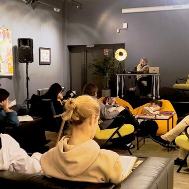 Reading Party w Bibliotece czyli Muzyka dla Książek — widok na salę, uczestnicy czytają, na scenie DJ Kacper Kapsa odtwarza muzykę