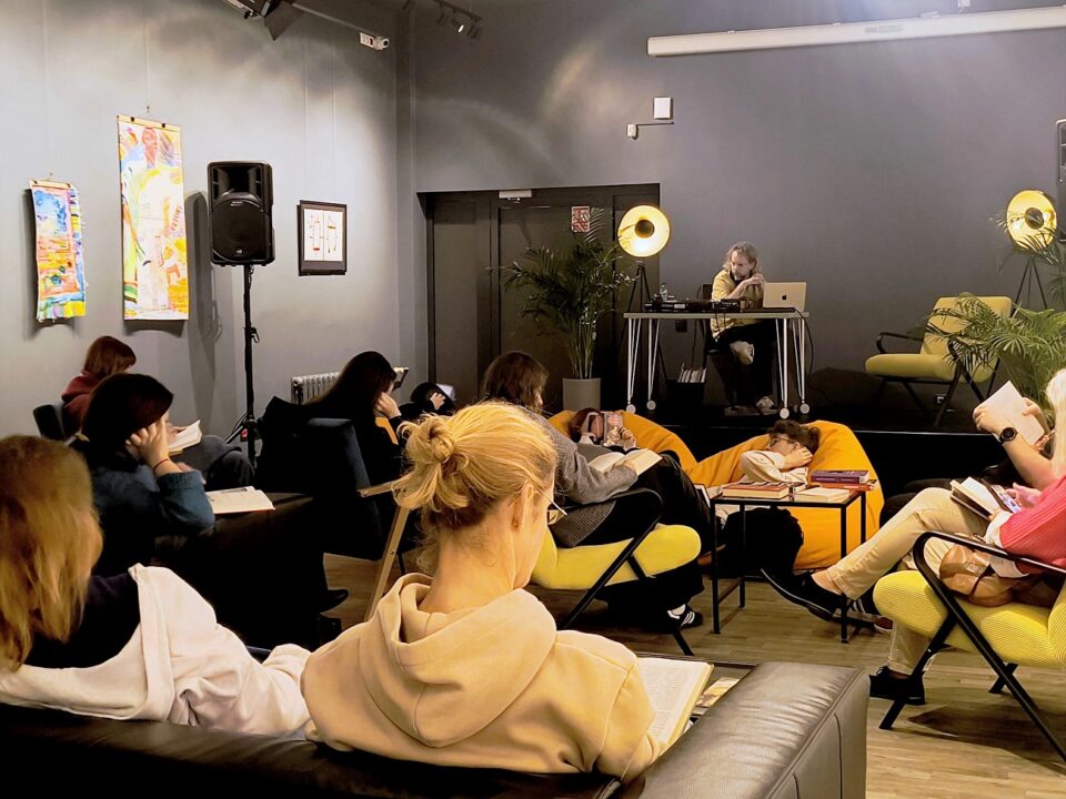 Reading Party w Bibliotece czyli Muzyka dla Książek — widok na salę, uczestnicy czytają, na scenie DJ Kacper Kapsa odtwarza muzykę