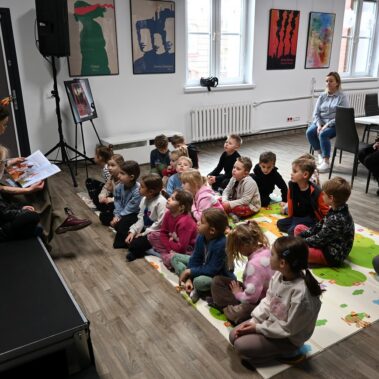 Dzieci siedzą na macie i słuchają co czyta bibliotekarka.