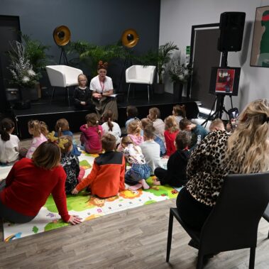 Dzieci siedzą na macie i słuchają co czyta bibliotekarka. Inne ujęcie
