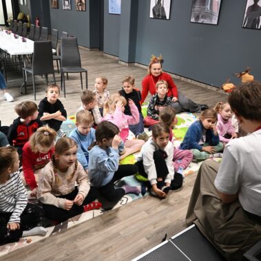 Dzieci siedzą na macie i słuchają co czyta bibliotekarka. Jeszcze inne ujęcie