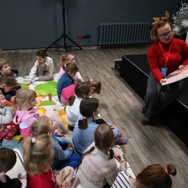 Druga bibliotekarka czyta książkę na głos, dzieci słuchają