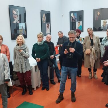 Goście wernisażu słuchają, w tle wiszą zdjęcia z wystawy