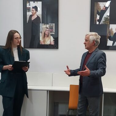 Prowadząca spotkanie rozmawia z Stanisławem Junkiertem, w tle wiszą zdjęcia z wystawy