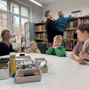 Bibliotekarka i dzieci rodzice. W tle ojciec pokazuje chłopcu zabawki leżące na regale. Na stole leżą koszyki do prac plastycznych