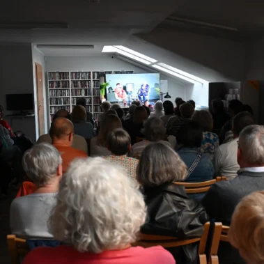 W Wypożyczalni Muzycznej goście słuchają bezpośredniej relacji na ekranie telewizora