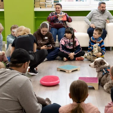 Dzieci siedzą na podłodze w kółko, w środku leży pies Lara