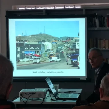 Na ekranie jest wyświetlany slajd — zdjęcie stolicy Grenlandii — Nuuk, wygląda skromnie bez wieżowców.