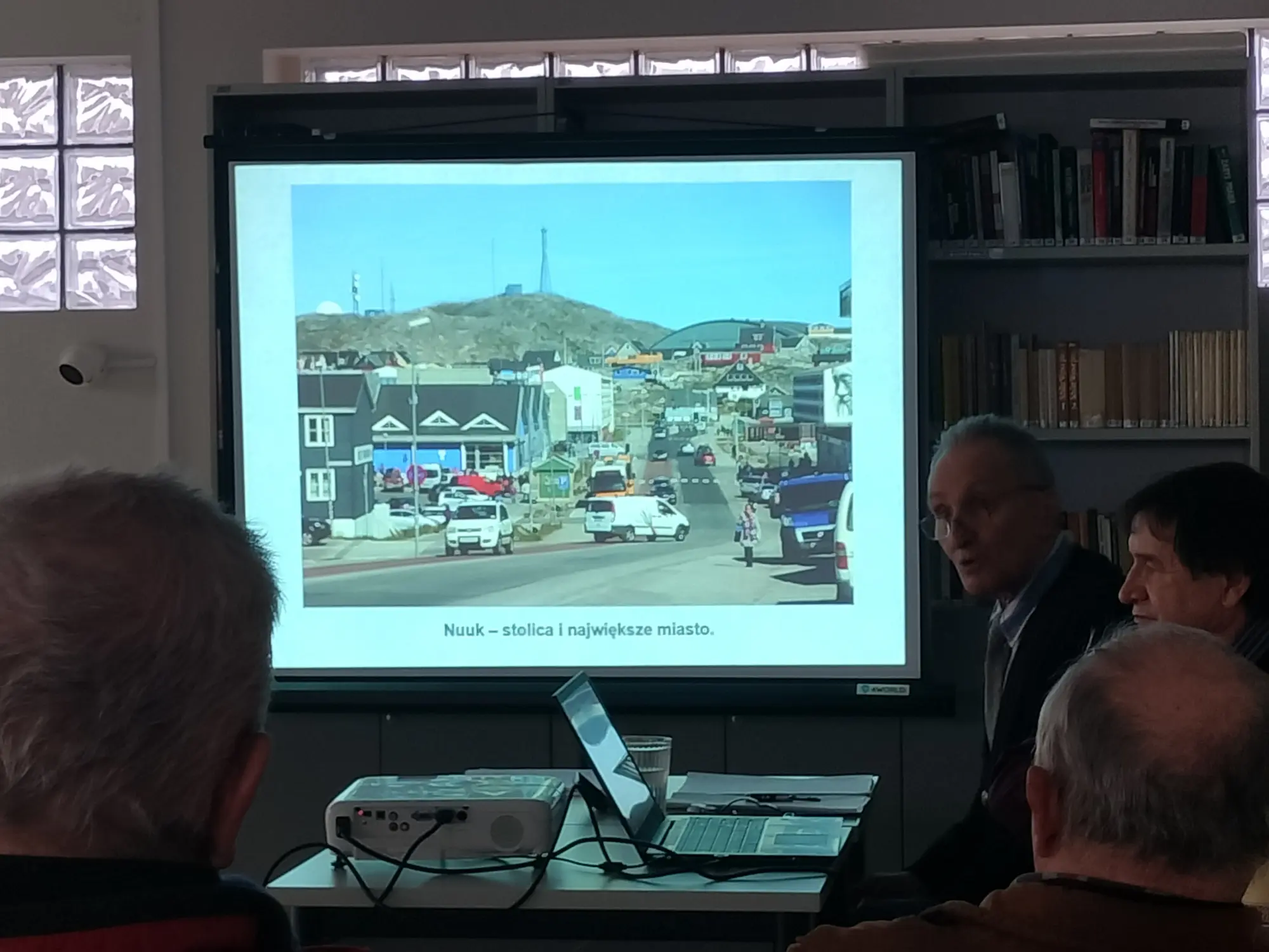 Na ekranie jest wyświetlany slajd — zdjęcie stolicy Grenlandii — Nuuk, wygląda skromnie bez wieżowców.
