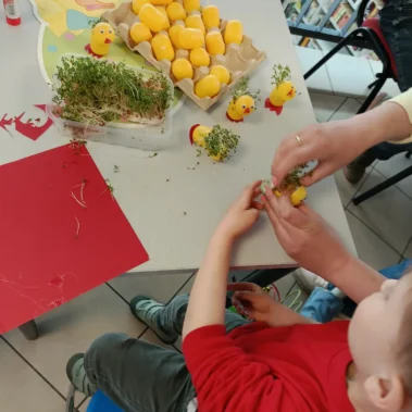 Z pomocą chłopiec wkładają zielska do jajka, na stole leżą rzeżuchy, puste jajka niespodzianki