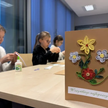 Przykładowa kartka wielkanocna z motywami kwiatowymi w technice quillingu, w tle uczestniczki przy pracy.
