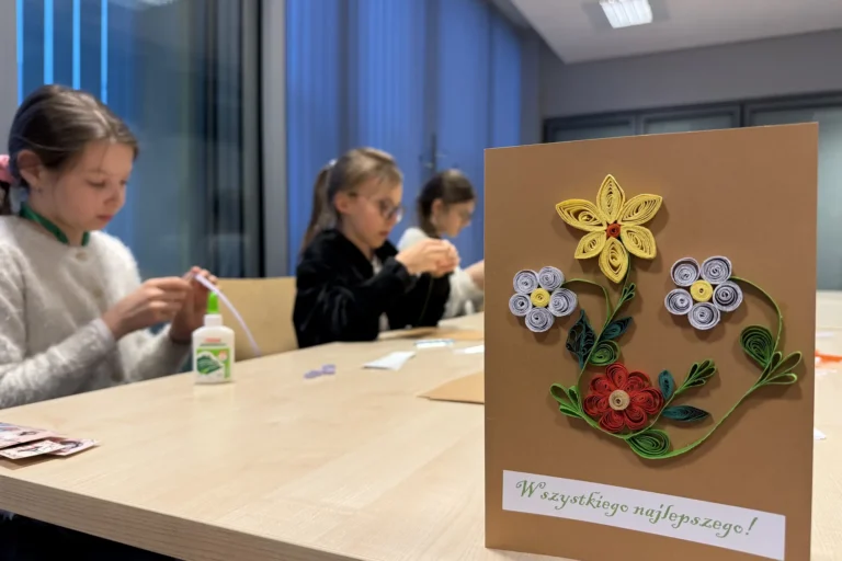 Przykładowa kartka wielkanocna z motywami kwiatowymi w technice quillingu, w tle uczestniczki przy pracy.