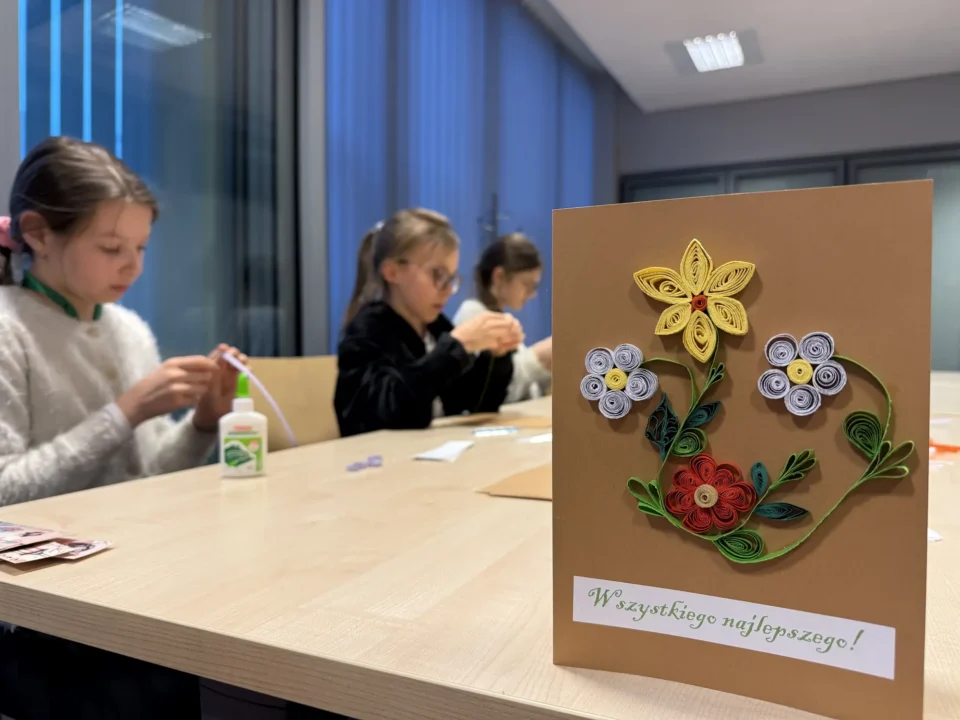 Przykładowa kartka wielkanocna z motywami kwiatowymi w technice quillingu, w tle uczestniczki przy pracy.
