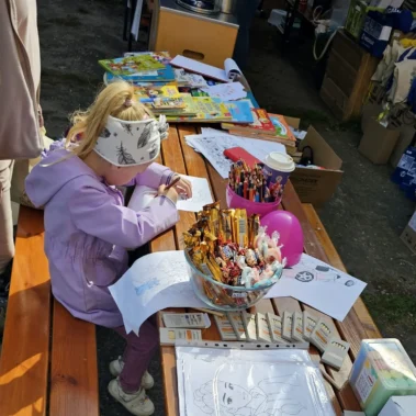 Dziewczynka rysuje, a stole leżą różne rysunki i przybory do rysowania oraz słodycze
